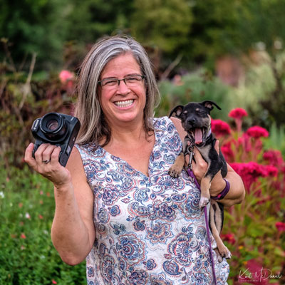 Woman smiling and holding a camera in one hand in a yawning puppy in the other. There is greenery and flowers behind them.